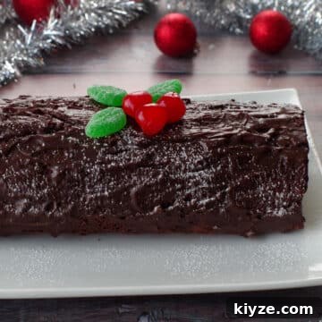whole black forest yule log cake on a white platter with tinsel and red tree decorations in background