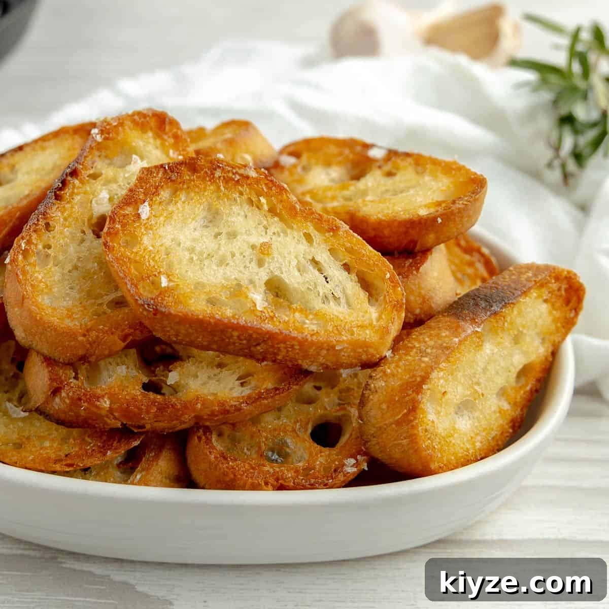 Crispy Garlic Crostini with Olive Oil and Herbs 20 Crostini in a white dish, garlic and rosemary in the background.