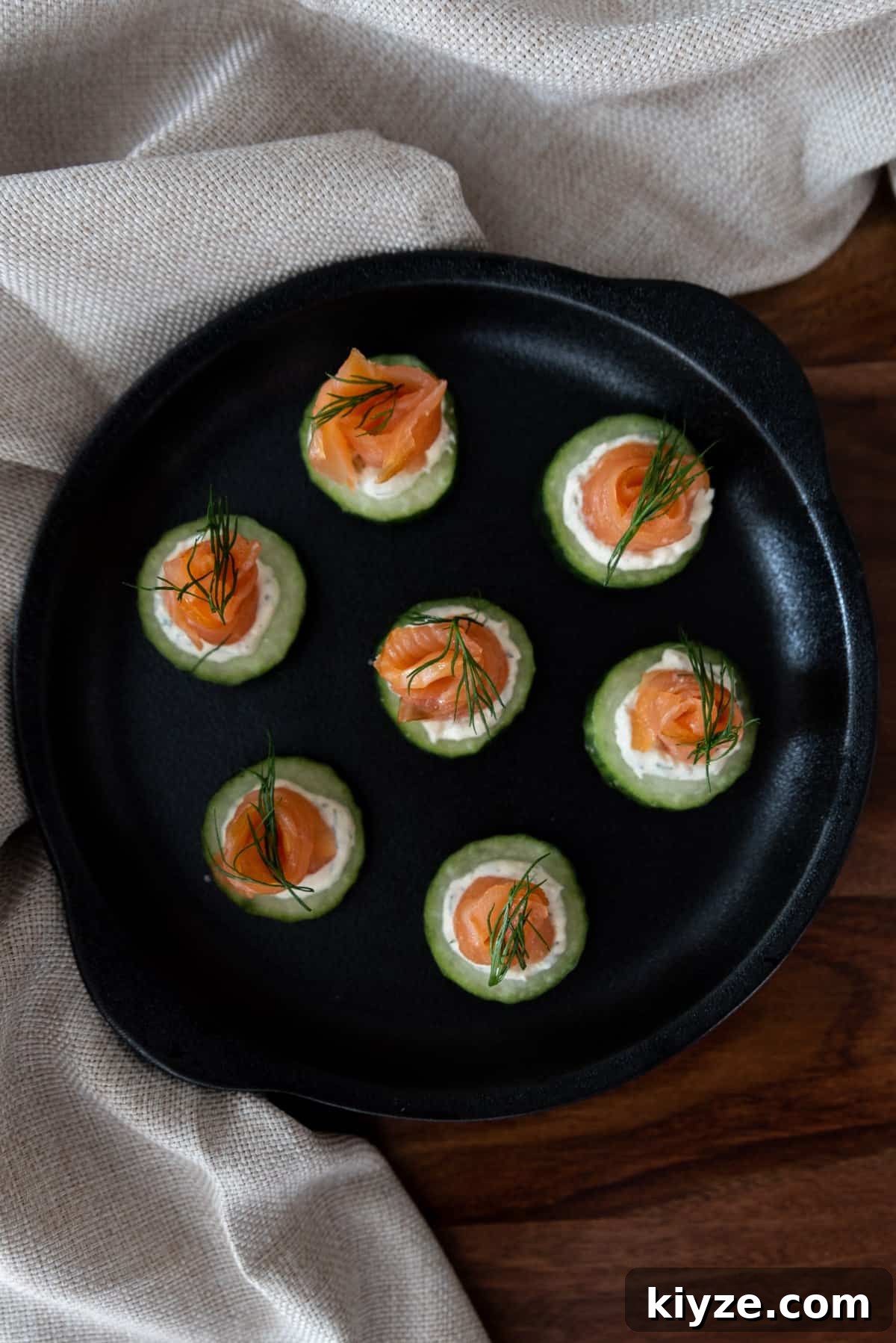 Cucumber Salmon Bites: Fresh Appetizer Recipe for Parties 6 cucumber bites in a round black cast iron dish, sitting on a timber table, with a beige napkin around the edge.
