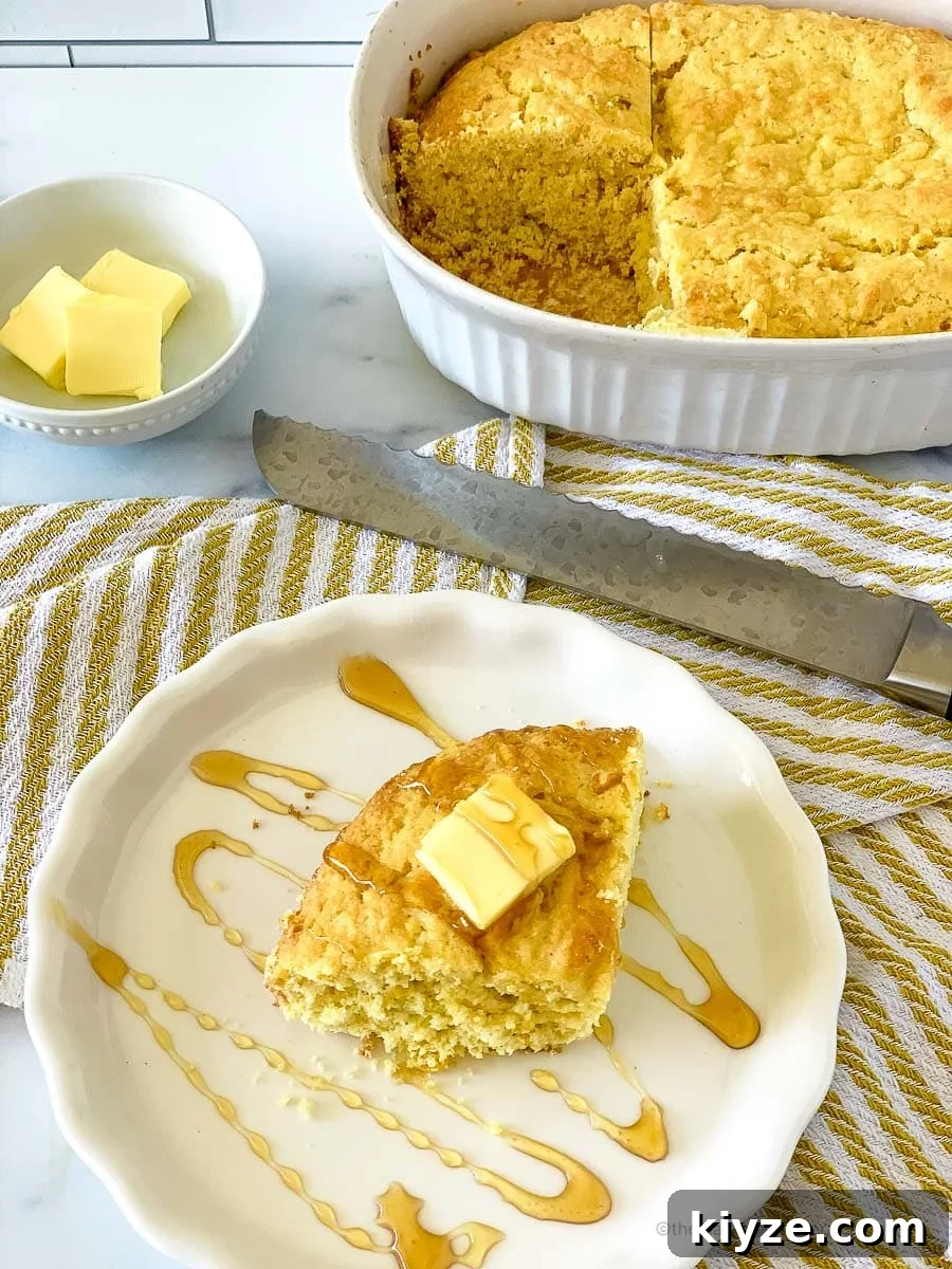 Jiffy Cornbread with Creamed Corn Casserole Recipe 2 A white plate with a slice of Jiffy cornbread with creamed corn with butter on top, and a casserole with cornbread, knife, and a small bowl with butter in the back.