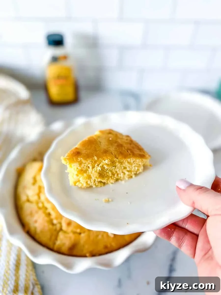 Jiffy Cornbread with Creamed Corn Casserole Recipe 3 a hand holding a white plate with a slice of Jiffy cornbread with a casserole dish of cornbread, yellow striped cloth, plate, and a bottle of honey in the background.