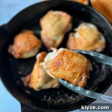 A golden brown chicken thigh is held with tongs in the foreground above a black cast iron pan of chicken thighs.