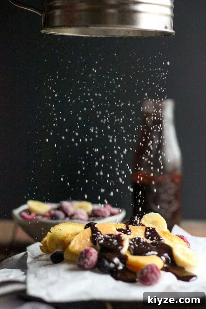 Gluten-Free Coconut Flour Crepes Recipe 4 Photo shows powdered sugar being sprinkled onto a plate of crepes with chocolate and berries