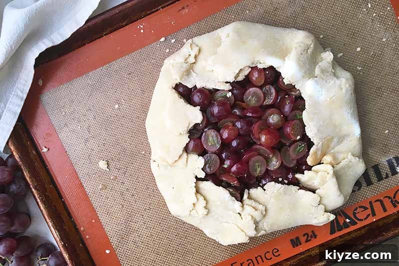 Grape Galette: Turn Fresh Grapes into a Rustic Dessert 13 Horizontal image of an unbaked open-faced grape pie on a sheet pan lined with a silicone mat.