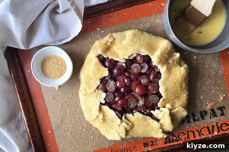 Grape Galette: Turn Fresh Grapes into a Rustic Dessert 14 Horizontal image of an unbaked galette with egg wash and sugar on a sheet pan lined with a silicone mat.