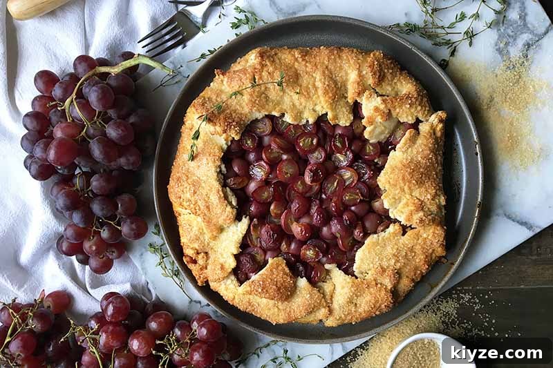 Grape Galette: Turn Fresh Grapes into a Rustic Dessert 17 Horizontal image of a whole baked galette on a marble board next to whole grapes thyme leaves, sugar, and a metal fork.