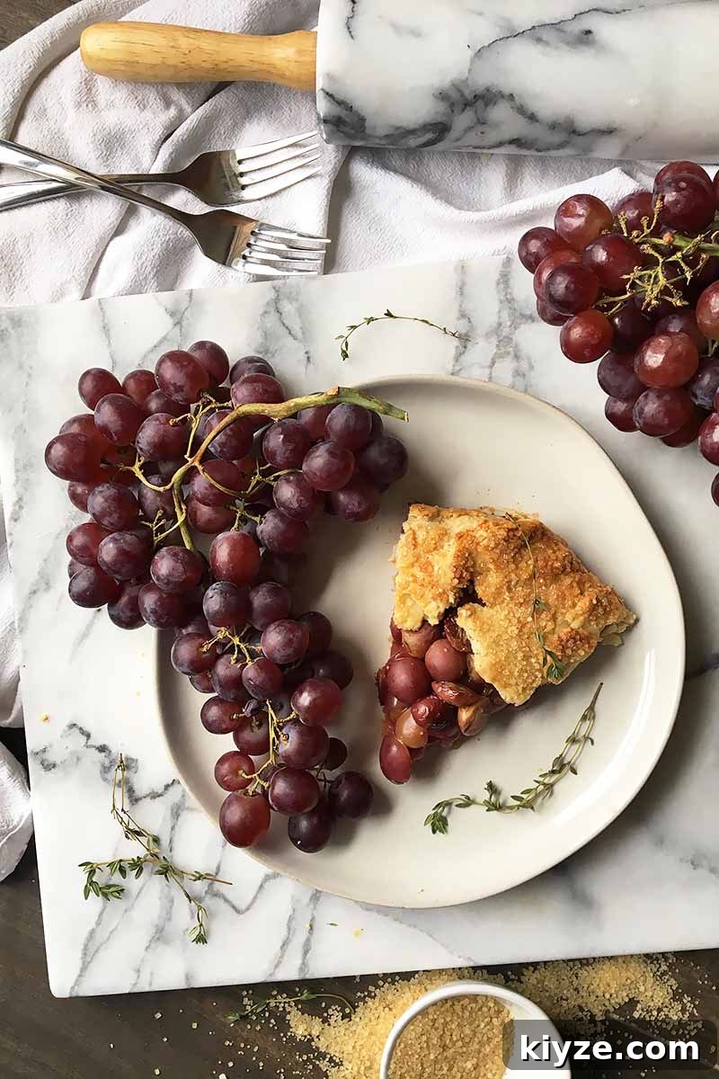 Grape Galette: Turn Fresh Grapes into a Rustic Dessert 4 Vertical top-down image of a slice of grape pie on a white plate with more fresh fruit and fresh thyme leaves next to forks, sugar, and a rolling pin.