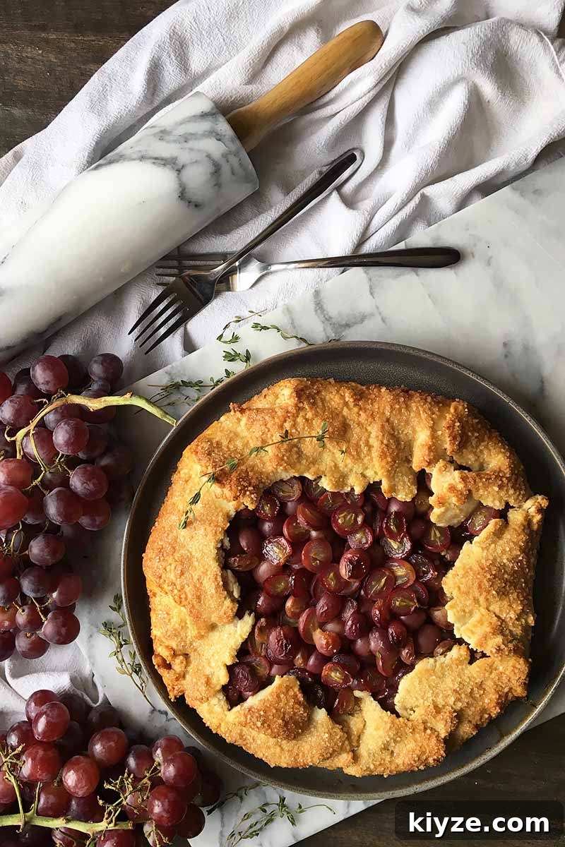 Grape Galette: Turn Fresh Grapes into a Rustic Dessert 6 Vertical image of a whole grape galette with thyme leaves on a marble slate, next to a marble rolling pin, a white towel, and forks.