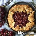 Horizontal image of a whole baked galette on a marble board next to whole grapes thyme leaves, sugar, and a metal fork.