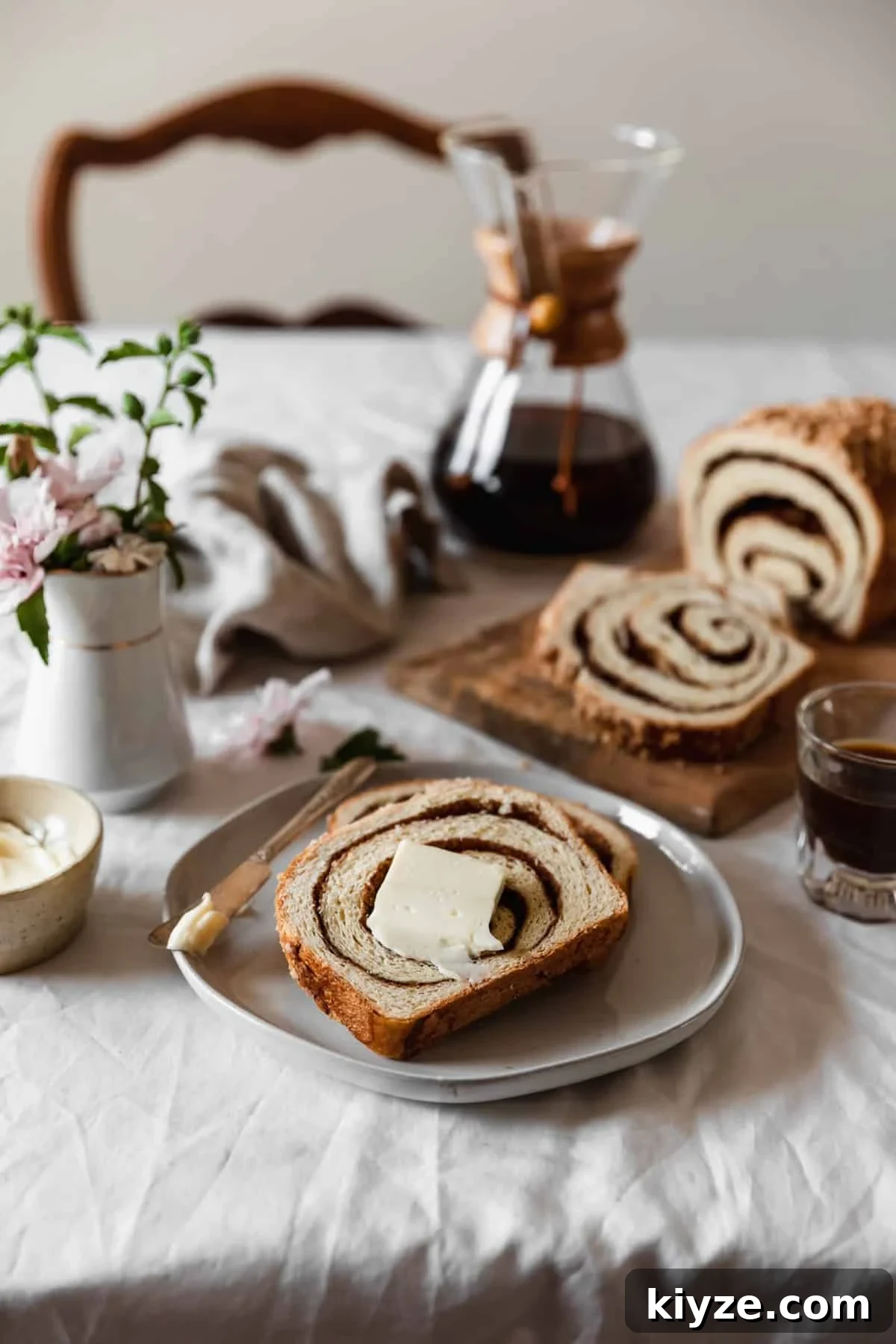 Cinnamon Swirl Bread with Crumbly Streusel Topping 2 A white plate with two slices of cinnamon swirl bread topped with a smear of butter next to a wood board with a loaf of bread, a white vase of pink flowers, a cream-colored bowl of butter, a Chemex, and a beige linen on a white table cloth with a wooden chair in the background.