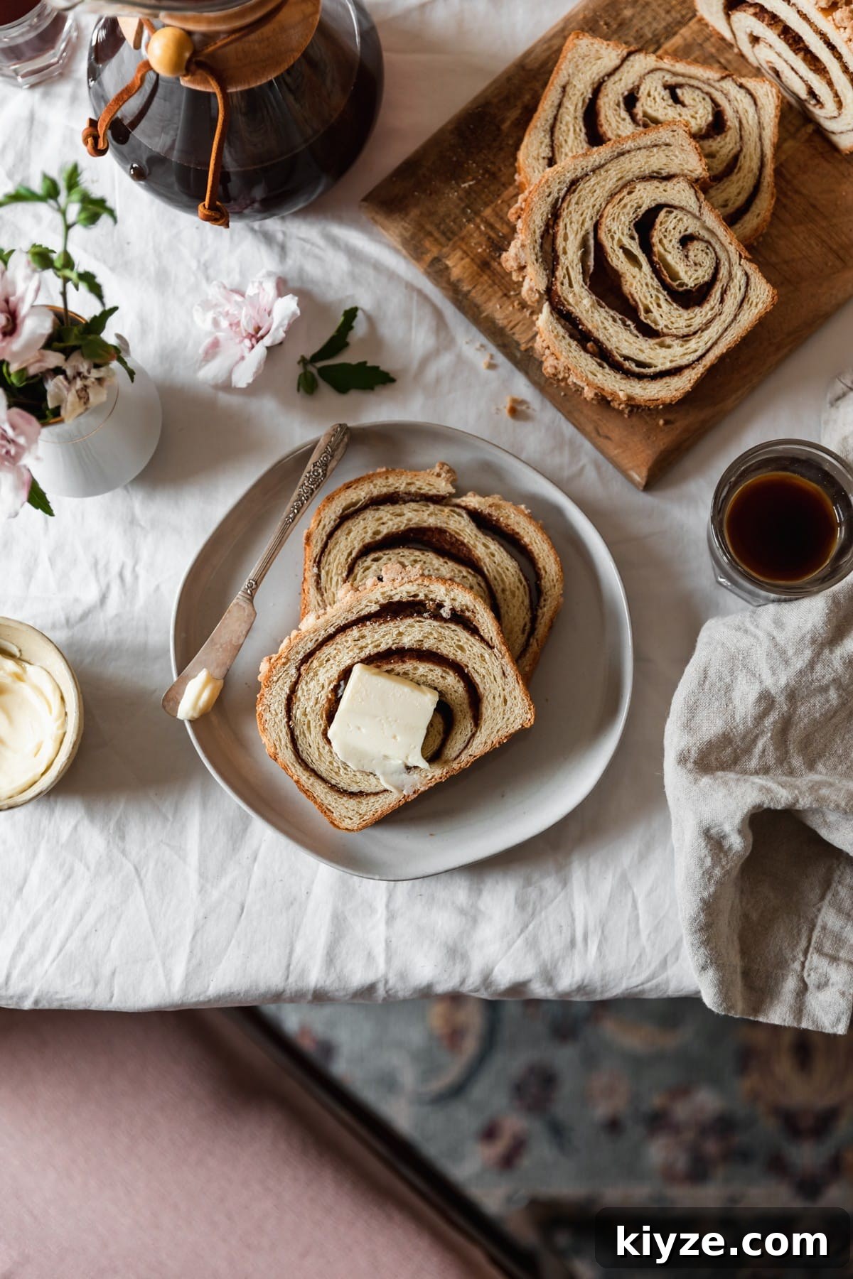 Cinnamon Swirl Bread with Crumbly Streusel Topping 10 An overhead image of two slices of cinnamon swirl bread with streusel and a butter knife on a white plate. The plate is next to a wood board in the right corner topped with more bread, a beige linen, cups of coffee, a Chemex, and a bowl of butter on a white table cloth. In the bottom of the photo, you can see a green and pink vintage rug and a pink seat cushion.