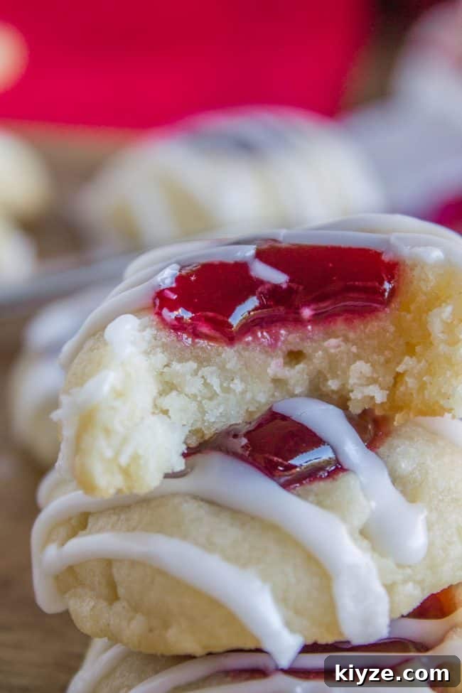 Almond Raspberry Thumbprint Cookies: Chewy Jam-Filled Recipe 4 Raspberry Thumbprint Cookies with icing in a stack, the top cookie with a bite missing.