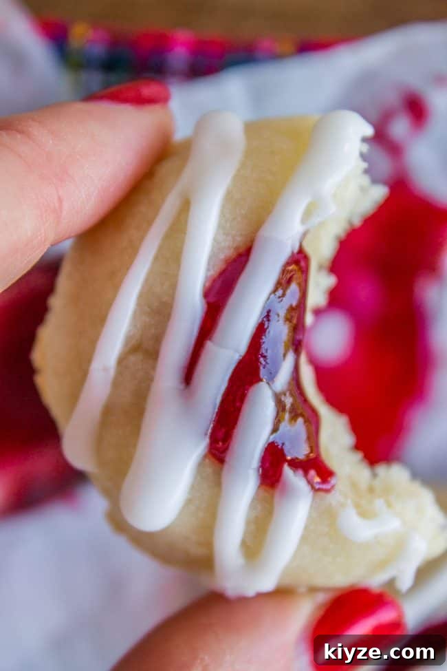 Almond Raspberry Thumbprint Cookies: Chewy Jam-Filled Recipe 6 Glaze-drizzled shortbread thumbprint cookie with bite taken, held by manicured hand.