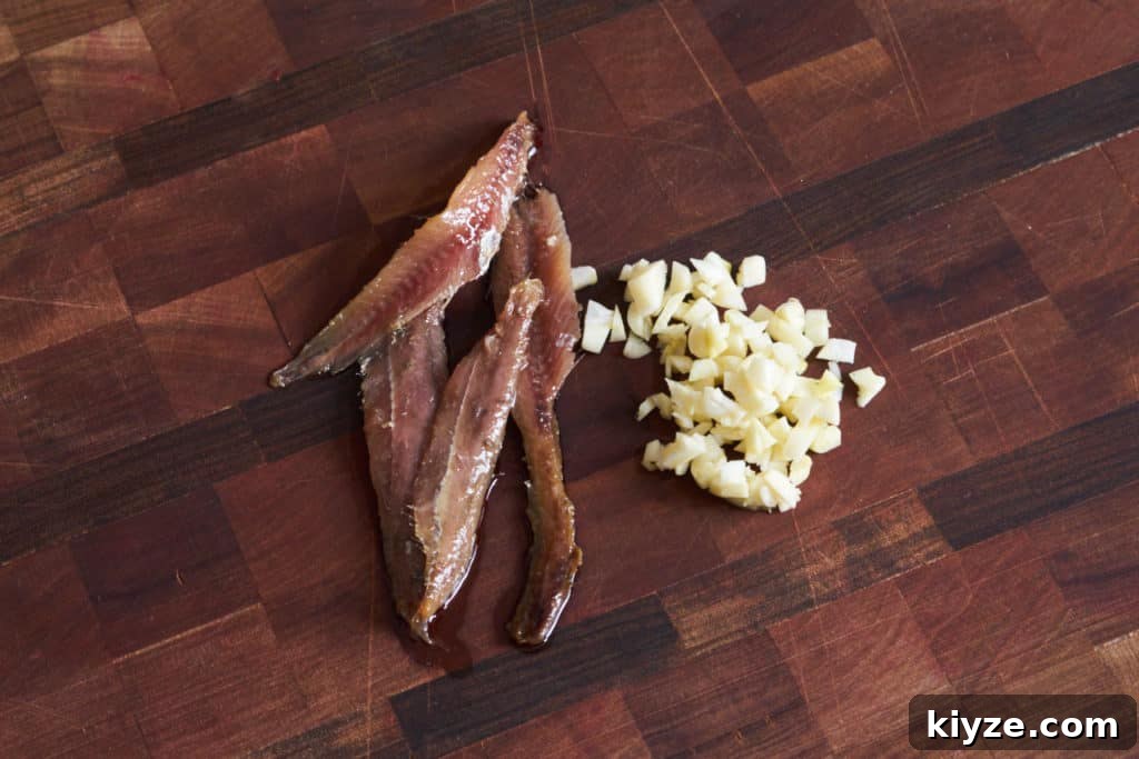Pasta with Green Olives, Capers & Fresh Herbs Recipe 5 Four oil-packed anchovies and chopped garlic on a wooden cutting board.