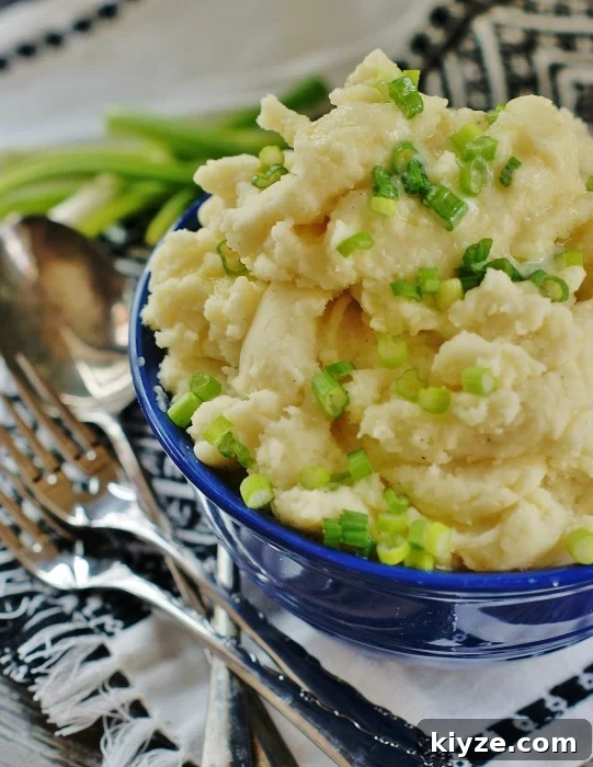 Upclose of a blue Fiestaware serving bowl piled high with horseradish mashed potatoes garnished with chives and drizzled with butter. A white and black tea towel, silverware and green onions are in the background.