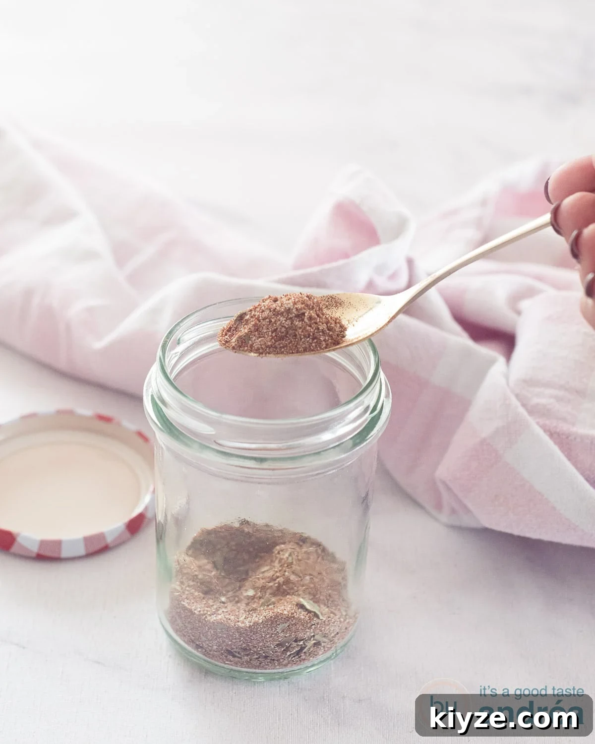 Homemade Chicken Seasoning Mix: Flavorful DIY Blend for Any Dish 2 A glass jar with chicken spices, a golden spoon scoops out a spoonful of homemade mix. A pink and white tea towel in the back.