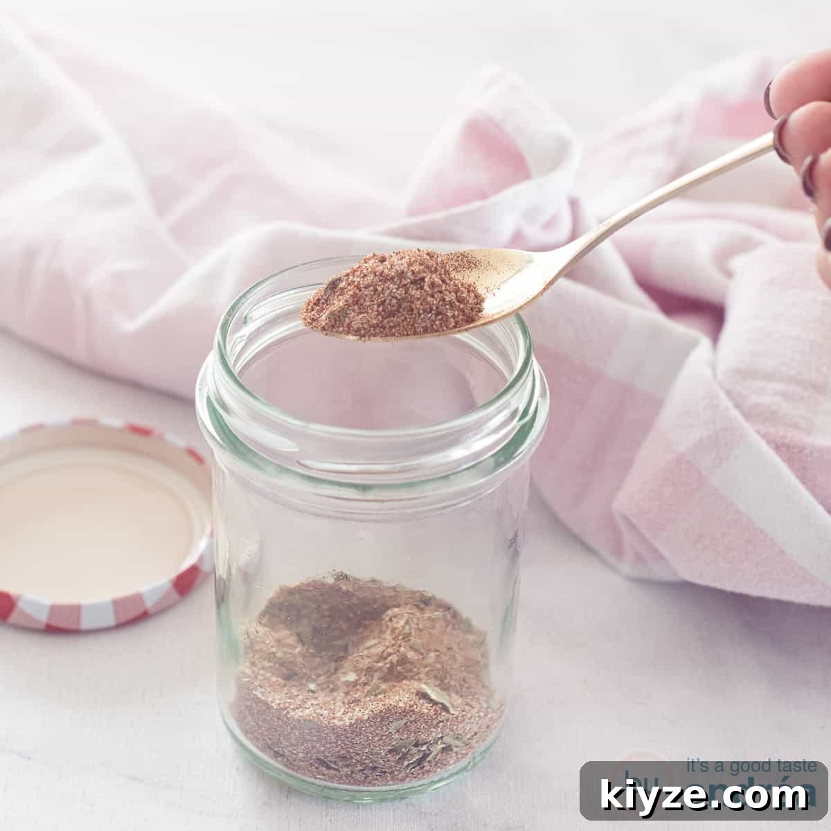 Homemade Chicken Seasoning Mix: Flavorful DIY Blend for Any Dish 4 A square photo with a glass jar with chicken spices that are mixed themselves. A spoonful of mix is scooped out. A pink white tea towel in the background.
