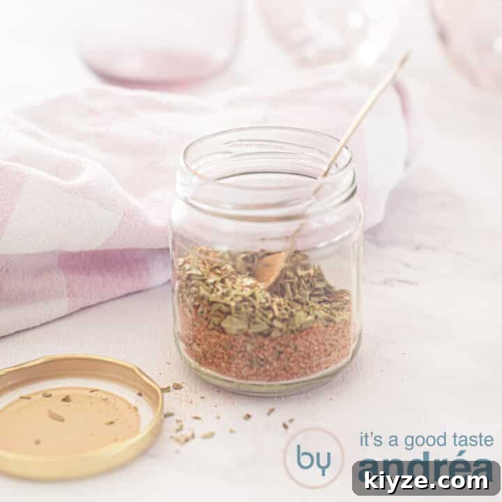 A square photo with a glass jar filled with gyros spice mix in the middle. A golden spoon is in it. The lid is next to it. A pink white tea towel and pink glasses in the background.