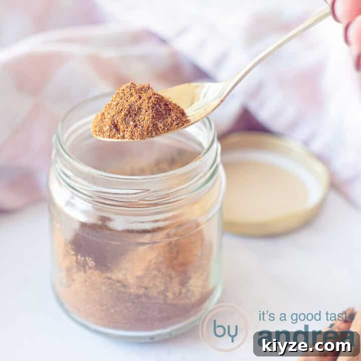 A square photo with a glass jar filled with shawarma spices. A pink white tea towel in the background. A spoon scoops the mix out of the jar.