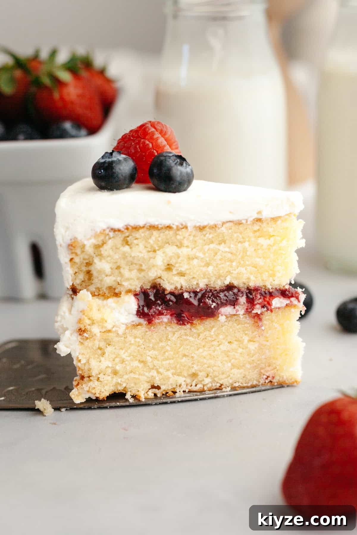 Mascarpone Whipped Cream Frosting Recipe — 5 Ingredients 8 A slice of vanilla sponge cake with white frosting, berry jam filling, and topped with blueberries and a raspberry.