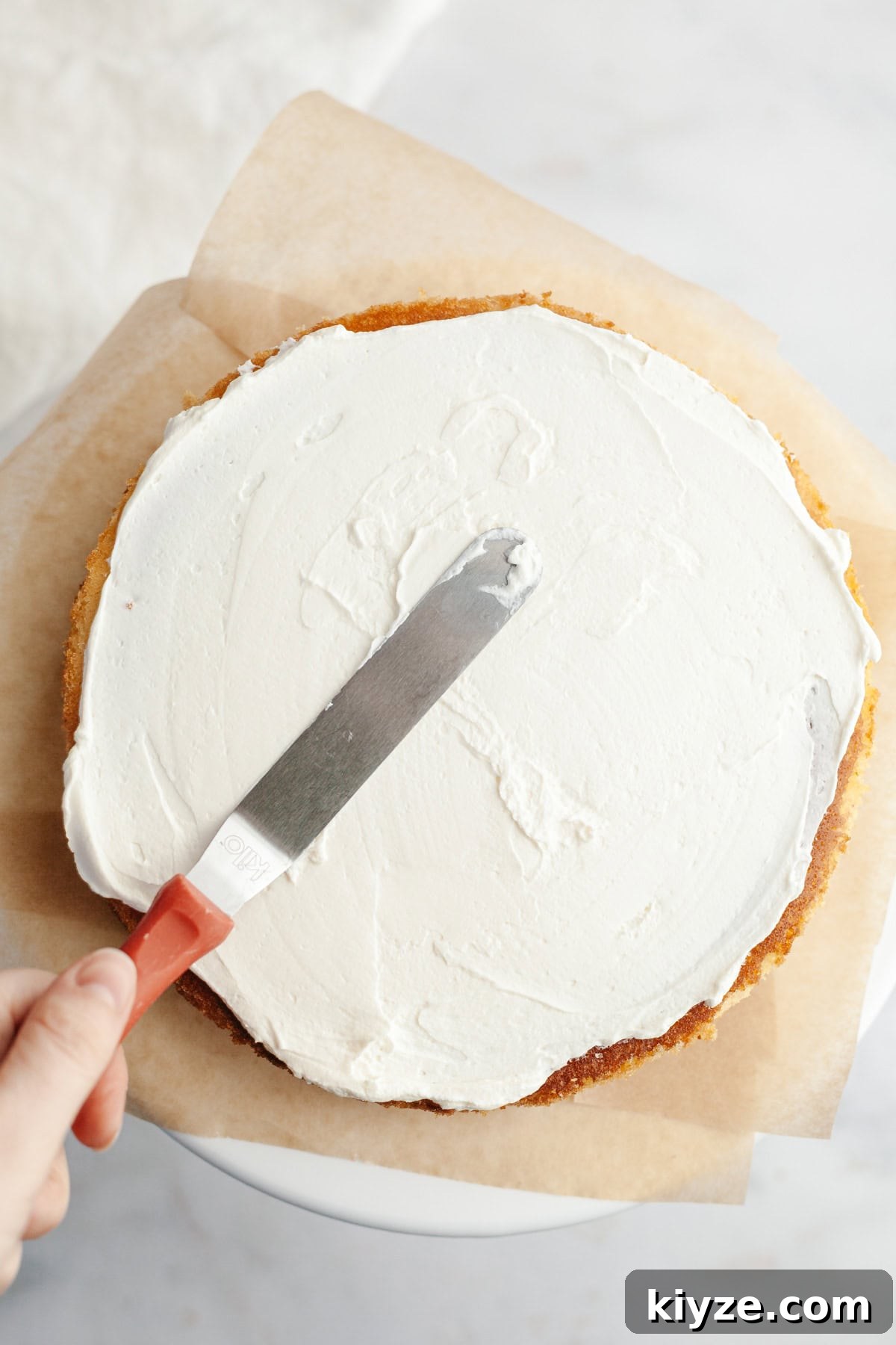 Mascarpone Whipped Cream Frosting Recipe — 5 Ingredients 9 A vanilla cake layer is on parchment paper, a hand is spread over whipped mascarpone frosting with a small offset spatula.