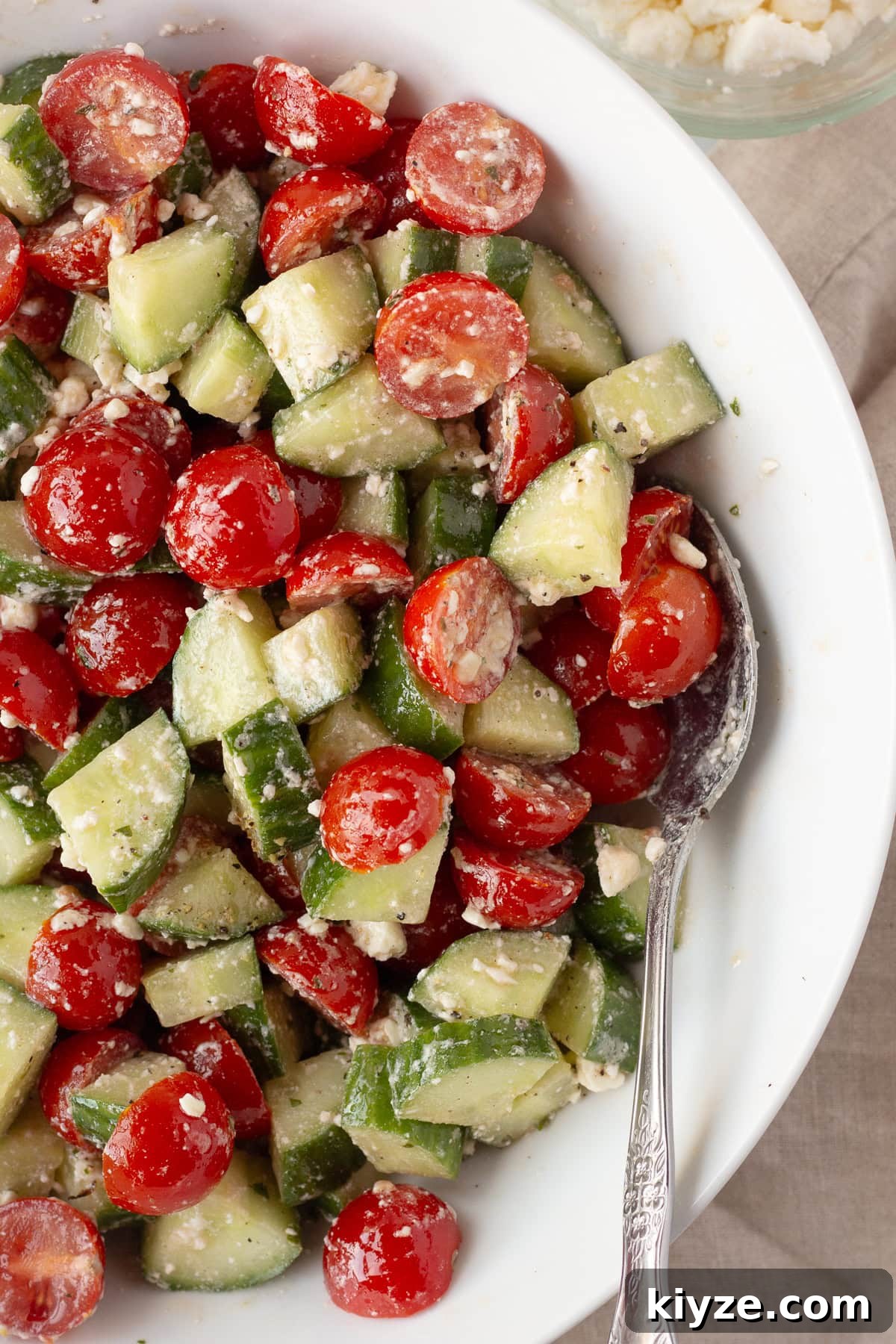Quick Cucumber Tomato Salad with Feta in 10 Minutes 2 A white bowl filled with a salad made of sliced cucumbers, halved cherry tomatoes, and crumbled feta cheese, with a metal spoon resting inside the bowl.