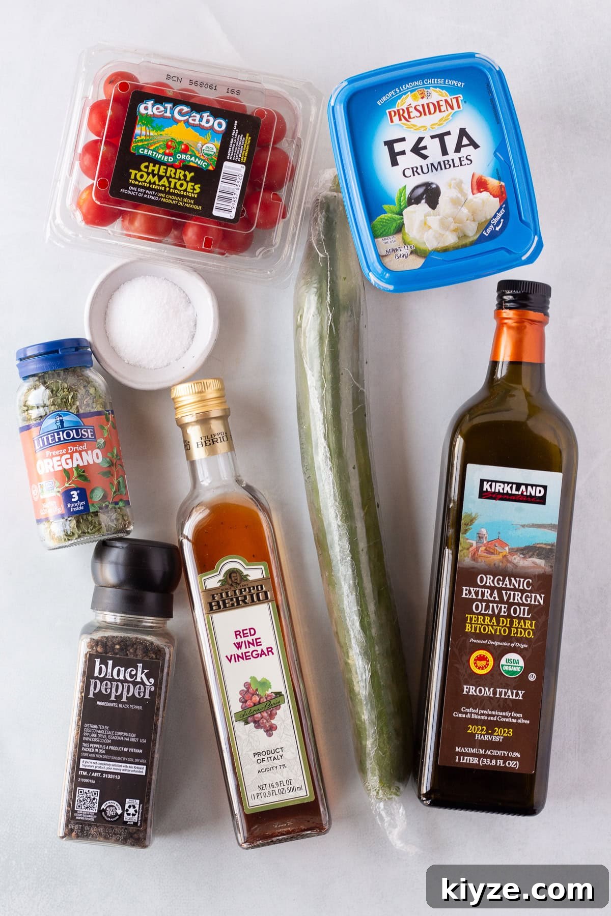 Quick Cucumber Tomato Salad with Feta in 10 Minutes 3 A container of cherry tomatoes, a tub of feta crumbles, a cucumber, a bottle of olive oil, a bottle of red wine vinegar, a jar of black pepper, a jar of oregano, and a small bowl of kosher salt on a white surface.