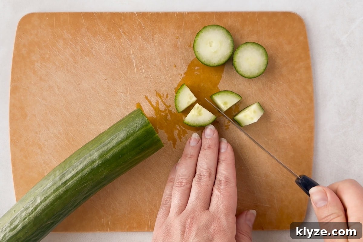 Quick Cucumber Tomato Salad with Feta in 10 Minutes 4 A hand is slicing a cucumber on a brown cutting board with a knife. Several round and quartered cucumber pieces are visible next to the partially cut cucumber.