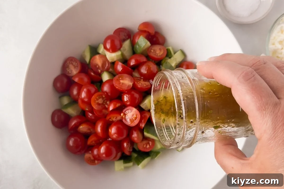Quick Cucumber Tomato Salad with Feta in 10 Minutes 7 A hand pours dressing from a glass jar over a bowl filled with sliced cherry tomatoes and chopped cucumbers.