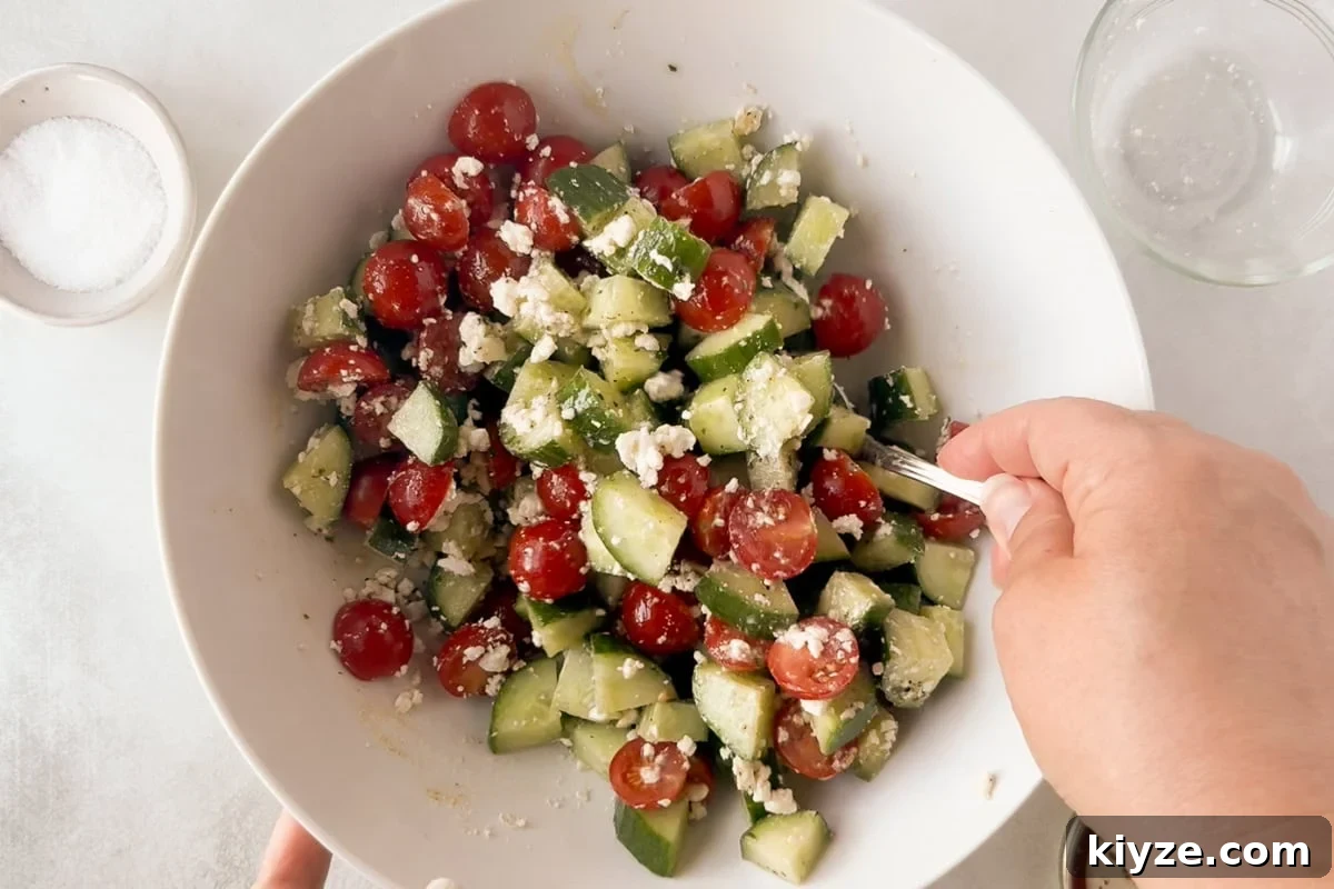 Quick Cucumber Tomato Salad with Feta in 10 Minutes 8 A hand holds a spoon, mixing a salad with chopped cucumbers, halved cherry tomatoes, and crumbled feta cheese in a white bowl.
