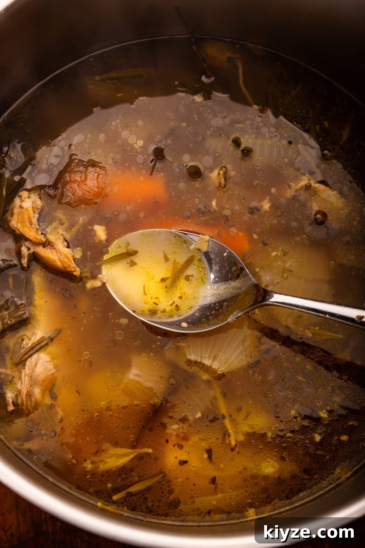 Homemade Chicken Stock from Leftover Roast Chicken 5 A spoon being dipped into a pot of chicken stock. The pot has vegetables, chicken pieces, and herbs in it.