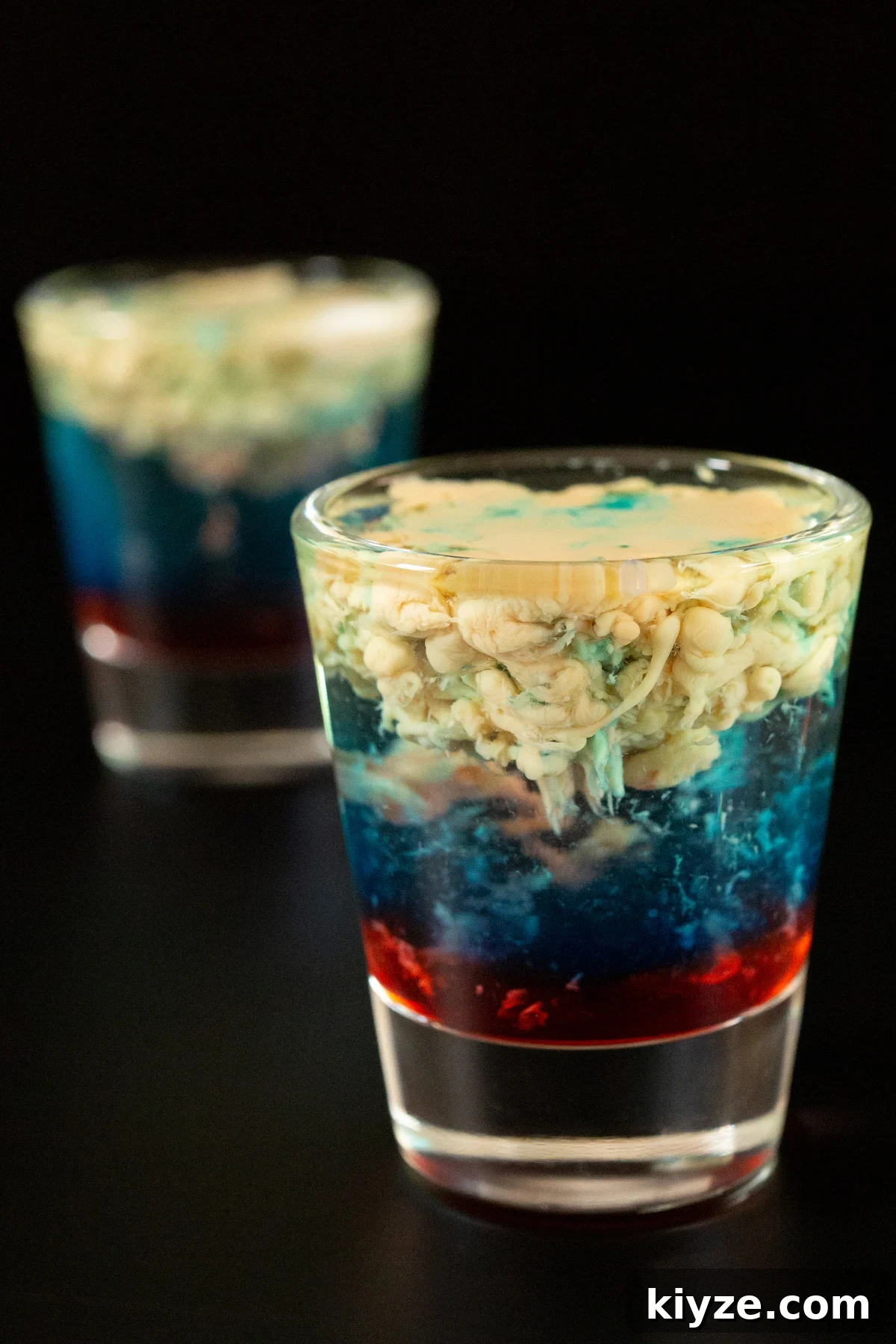 Alien Brain Hemorrhage Shot Recipe: How to Make the Layered Party Shooter 4 Two shot glasses on a black background. Each is filled with a layered shot that's red on the bottom, blue in the middle, and the top is a light brown liquid that has formed a brain-like texture.