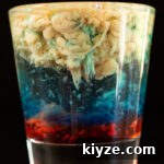 Alien Brain Hemorrhage Shot Recipe: How to Make the Layered Party Shooter 5 A shot glass is filled with a layered shot that is red on the bottom, then blue, then clear, and a light brown brain texture for the top layer.
