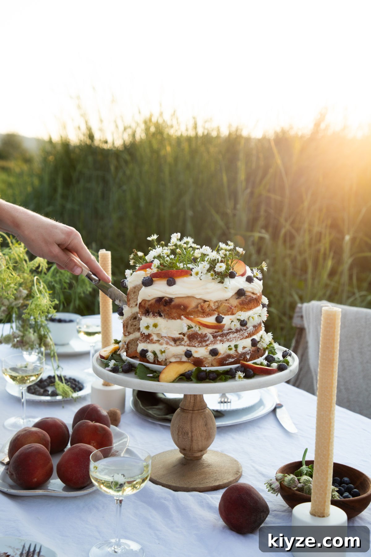 Homemade Peach Cake with Fresh Peach Glaze 3 a woman's hand cutting into a cake topped with peaches, blueberries, and camomile flowers on a white cloth covered table in a field