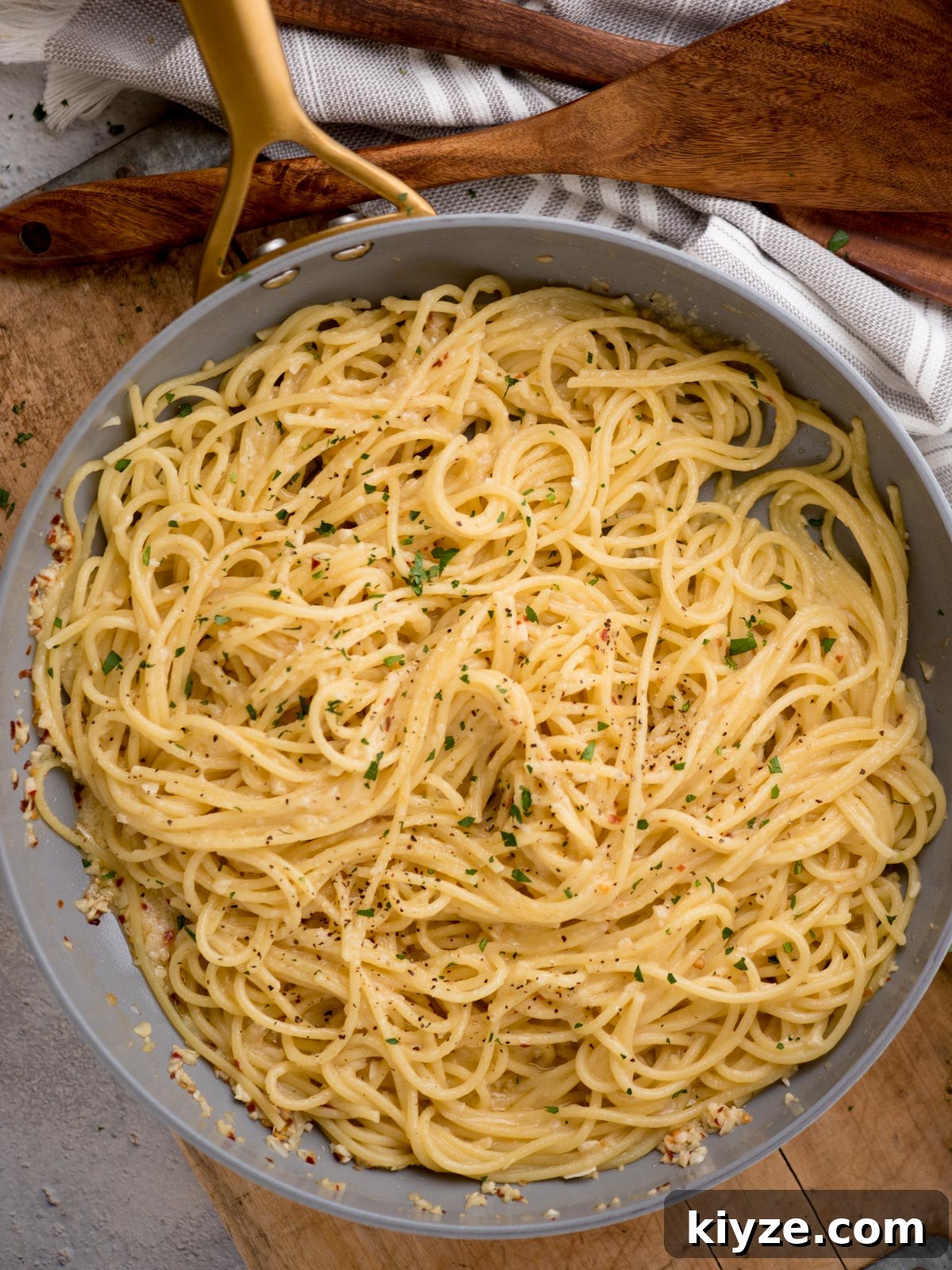 Creamy Lemon Garlic Pasta Recipe with Parmesan and Herbs 2 A skillet filled with freshly cooked spaghetti, lightly coated in a creamy sauce with a sprinkle of herbs and black pepper on top. A wooden spatula and a striped cloth are visible in the background.