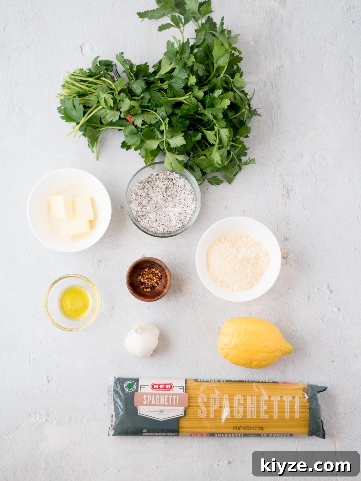 Creamy Lemon Garlic Pasta Recipe with Parmesan and Herbs 3 A bundle of spaghetti, fresh parsley, lemon, garlic bulb, a small bowl of parmesan, a small bowl of butter cubes, a small wooden bowl of red pepper flakes, and a small glass bowl of olive oil are arranged on a light surface.