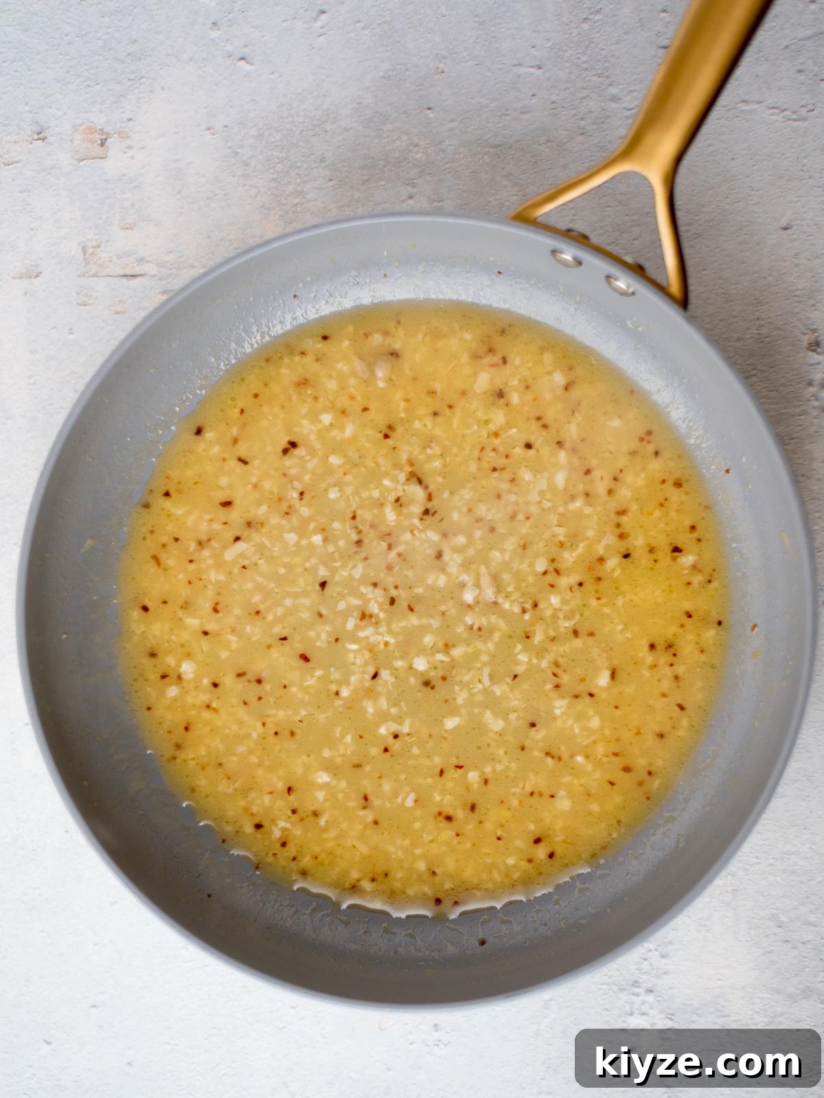 Creamy Lemon Garlic Pasta Recipe with Parmesan and Herbs 4 A frying pan with a gray interior and a gold handle contains a creamy, speckled sauce. The sauce appears to be a butter, garlic, and mustard mixture, with visible small pieces of garlic or seasonings dispersed throughout. The background is light gray.