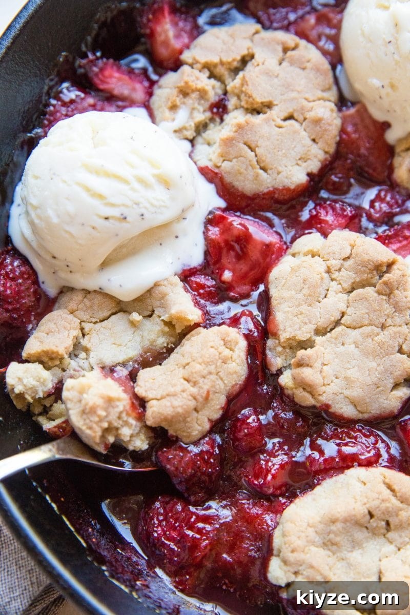 Vegan Gluten-Free Strawberry Cobbler Recipe for Summer Baking 8 Strawberry cobbler in a large cast iron skillet with scoops of melted ice cream on top and a spoon scooping out some of the cobbler.