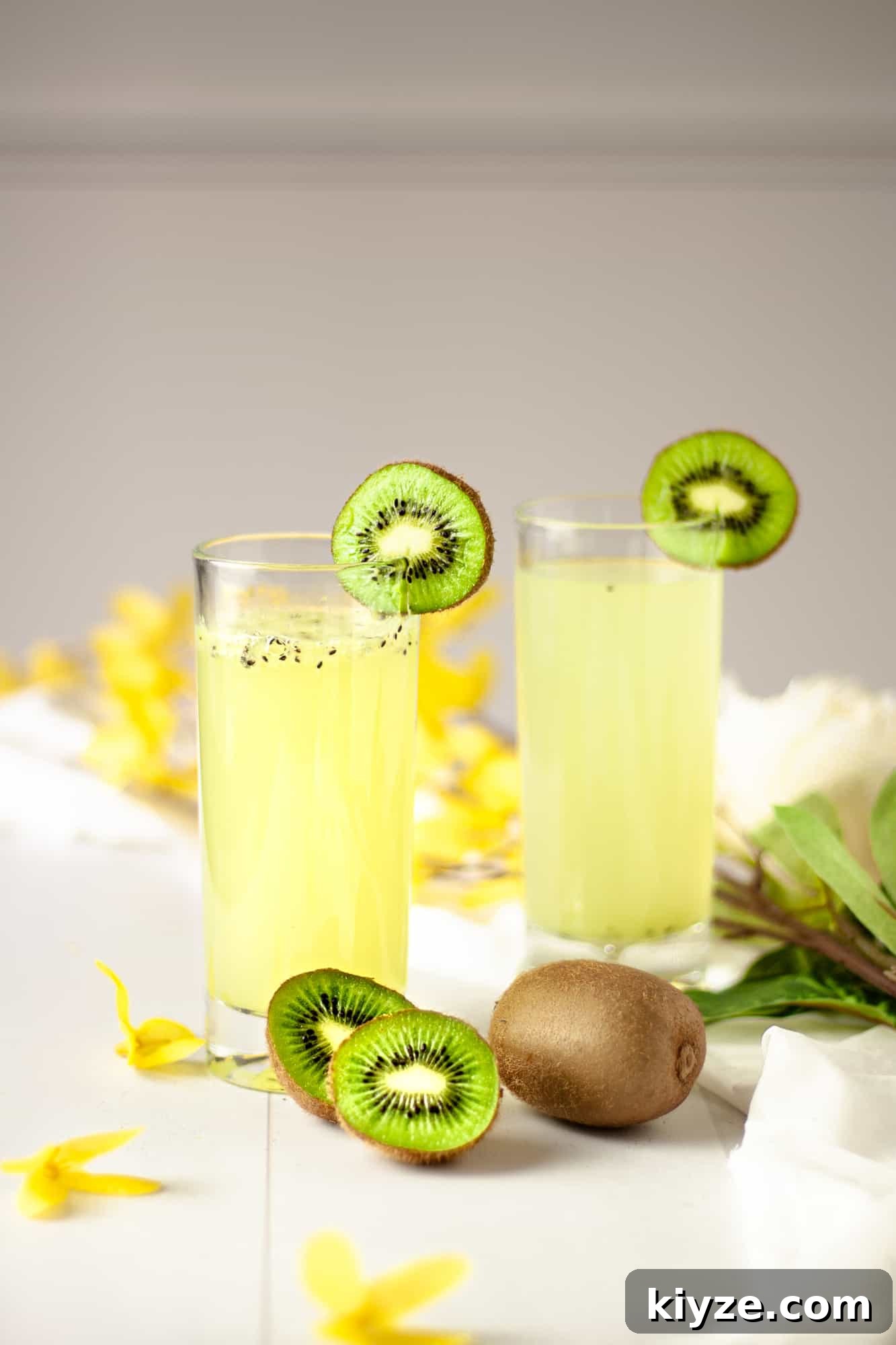 Refreshing Kiwi Gin Cocktail Recipe for Summer Sips 2 Two kiwi gin cocktails garnished with kiwi slices and sitting in a table with scattered yellow flowers.