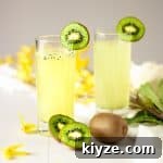 Two kiwi gin cocktails garnished with kiwi slices and sitting in a table with scattered yellow flowers.