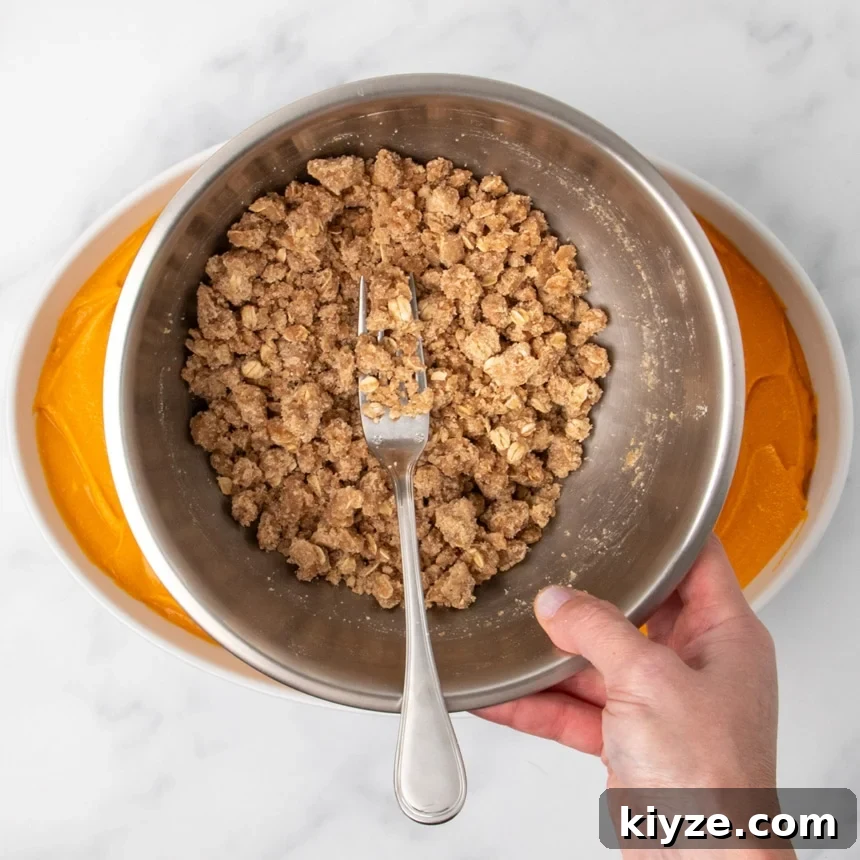Gluten-Free Sweet Potato Casserole Recipe for Holidays 12 A hand holding a metal bowl with a fork in a crumbly oat topping mixture above a dish filled with orange sweet potato filling.