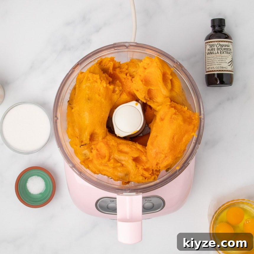 Gluten-Free Sweet Potato Casserole Recipe for Holidays 13 A food processor filled with cooked orange sweet potato pieces surrounded by small bowls of sugar, salt, 4 eggs out of the shell, and a bottle of vanilla extract on a white marble surface.