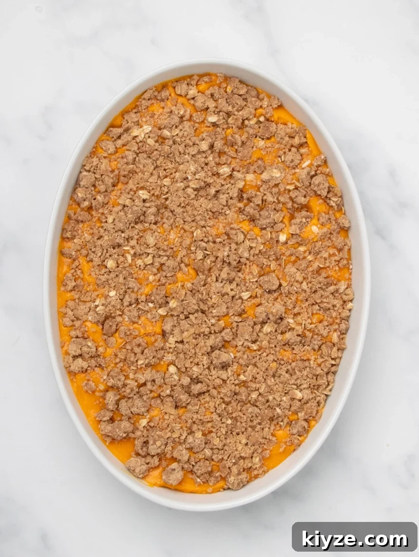 Gluten-Free Sweet Potato Casserole Recipe for Holidays 17 A white oval baking dish filled with orange sweet potato filling topped with an even layer of raw crumb mixture on a white marble surface.