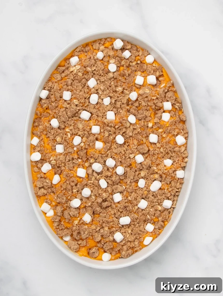 Gluten-Free Sweet Potato Casserole Recipe for Holidays 18 A white oval baking dish filled with orange sweet potato filling topped with an even layer of raw crumb mixture and scattered small white marshmallows, unbaked.