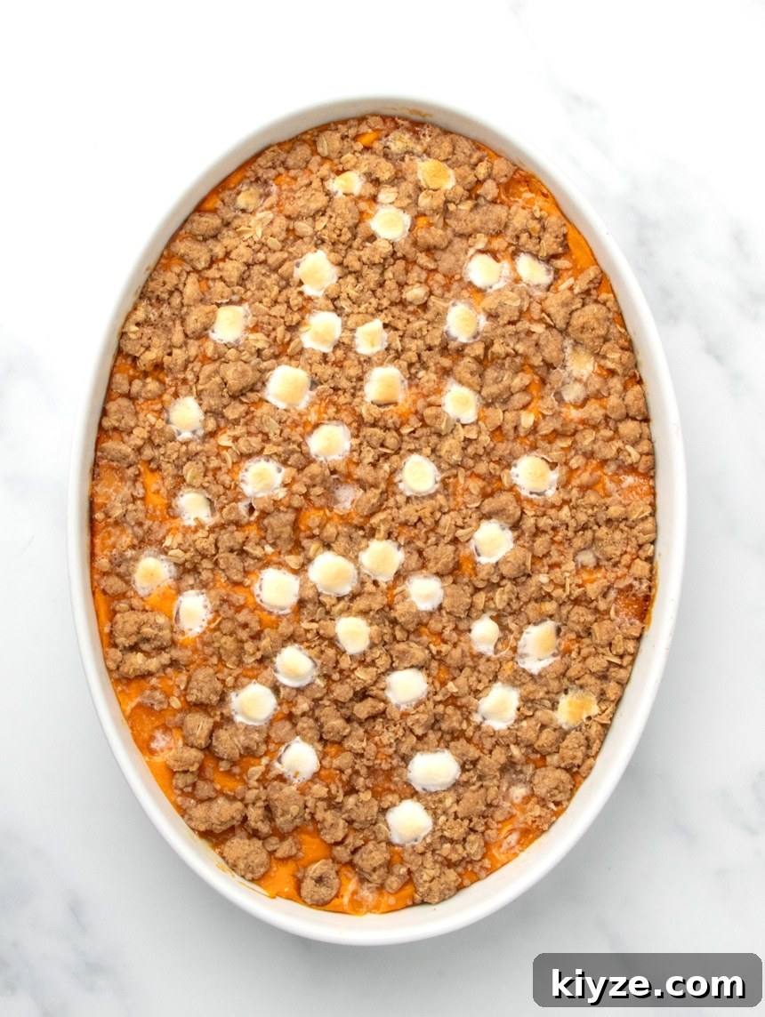 Gluten-Free Sweet Potato Casserole Recipe for Holidays 19 A white oval baking dish filled with orange sweet potato filling topped with a browned baked crumb mixture and melted marshmallows on a white marble surface.