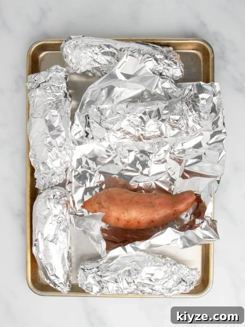 Gluten-Free Sweet Potato Casserole Recipe for Holidays 7 A metal baking sheet holding several sweet potatoes fully wrapped in aluminum foil, with one sweet potato uncovered in the center of the batch.
