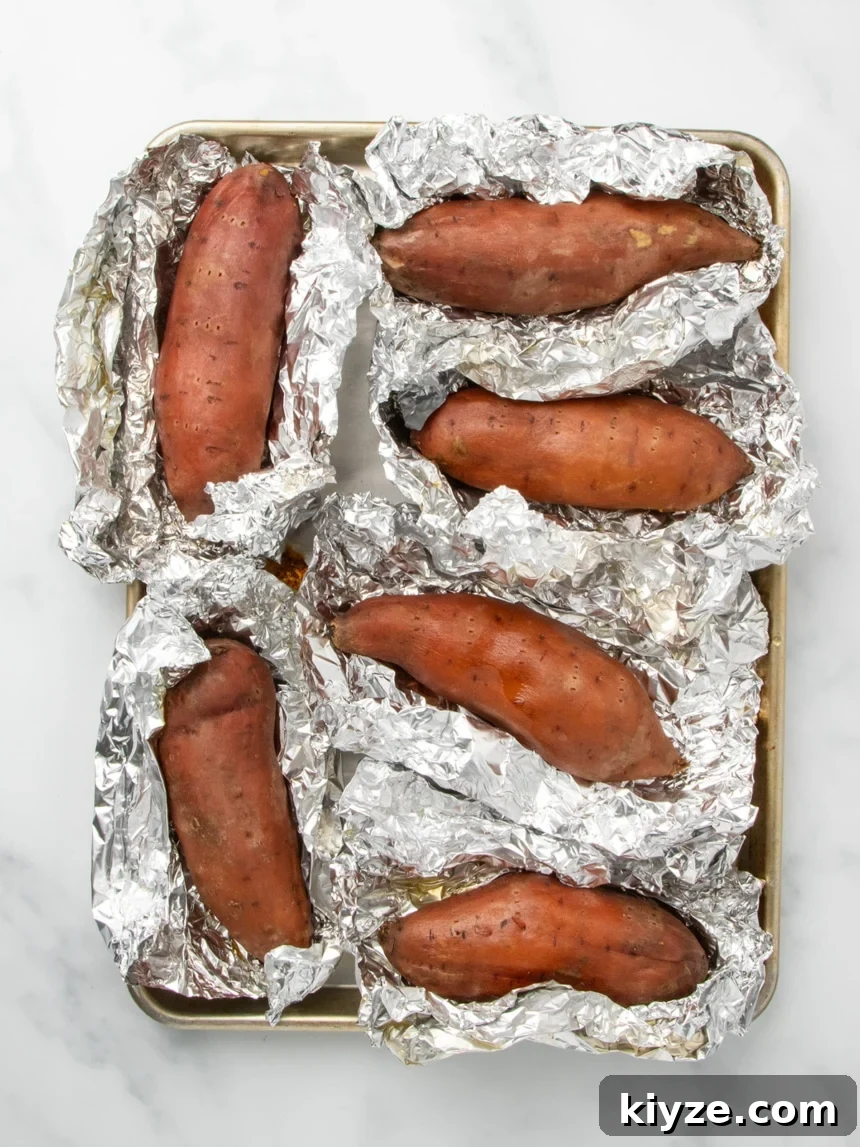Gluten-Free Sweet Potato Casserole Recipe for Holidays 8 A metal baking sheet holding six cooked sweet potatoes, each resting in partially opened aluminum foil.