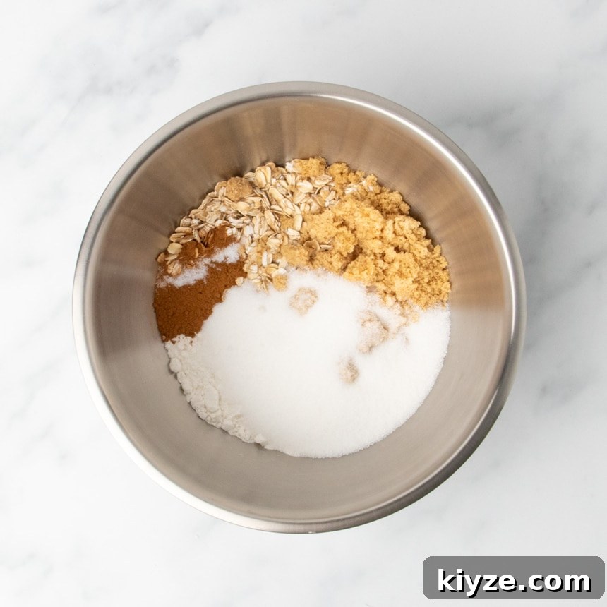 Gluten-Free Sweet Potato Casserole Recipe for Holidays 9 A metal mixing bowl containing dry ingredients including oats, brown sugar, white sugar, gluten free flour, and cinnamon on a white marble surface.