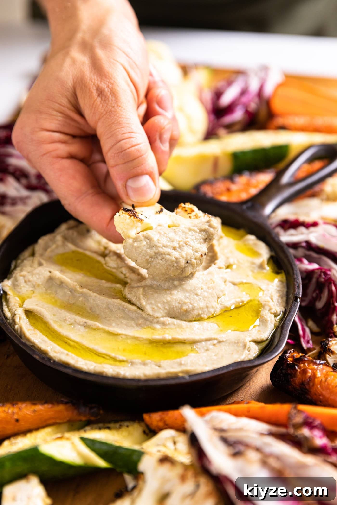 Authentic Baba Ganoush Recipe: Smoky Eggplant Dip Guide 3 Hand holding piece of grilled cauliflower dipping into light brown colored Baba ganoush drizzled on top with olive oil and sitting in black cast iron skillet
