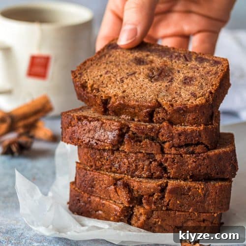 Five fruit cake loaf slices stacke on top of each other, there is a hand holding the top slice. In the background is a cup of tea and various whole spices.
